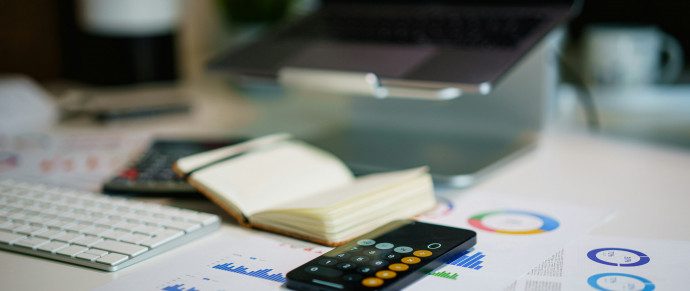 A desk with a laptop, keyboard, calculator, open notebook and printed charts