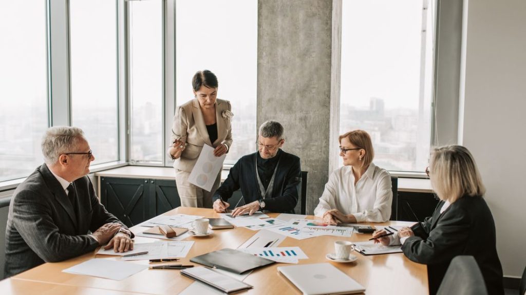 Senior professionals discussing documents around a boardroom table with a city view behind them